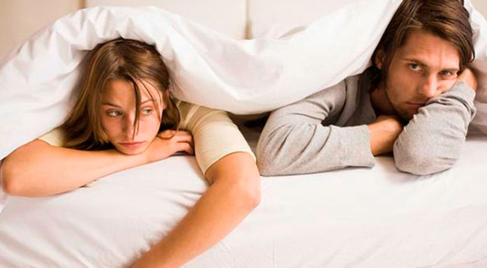 Jovem casal deitado de bruços na cama, com o lençol sobre o corpo, com aspecto triste